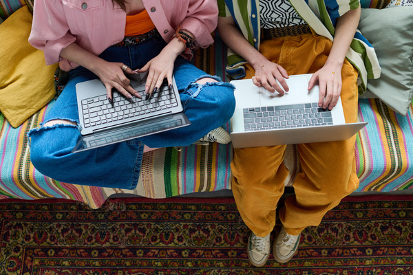 A girl in a pink jacket and jeans with holes uses a laptop sitting on a sofa with a striped plaid and pillows in a lotus position next to a friend in yellow pants typing in her laptop